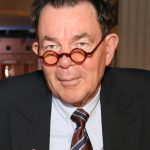 headshot of a man wearing a blazer over a collared shirt and tie. He is also wearing round-rimmed glasses.
